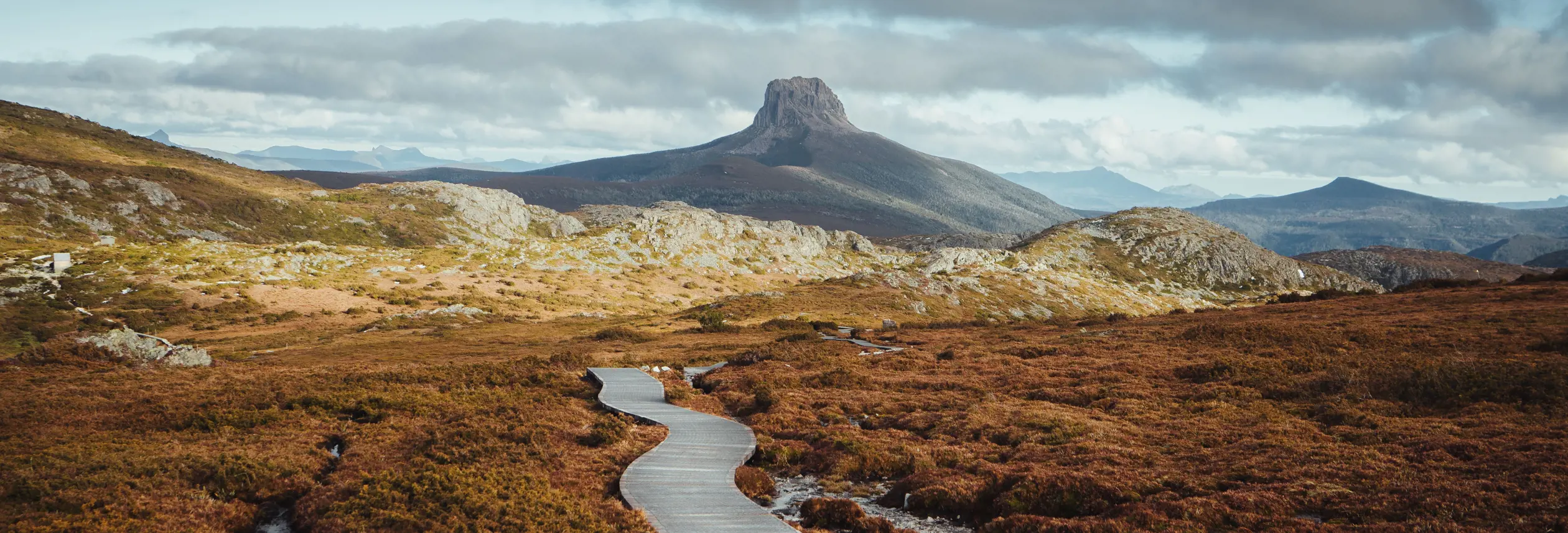 A boardwalk winds through alpine moorland toward a distinctive rocky peak on the Overland Track, Cradle Mountain