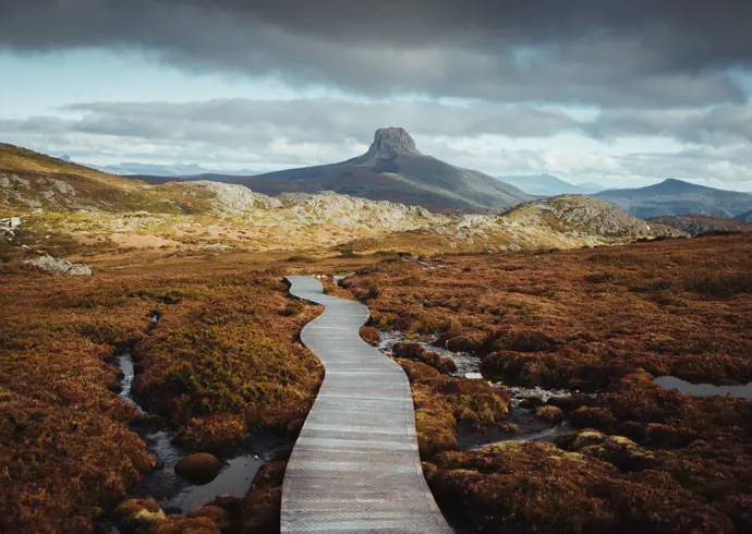 A boardwalk winds through alpine moorland toward a distinctive rocky peak on the Overland Track, Cradle Mountain