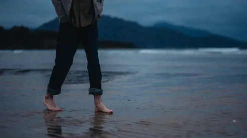 A person walks barefoot across wet sand on Sandpiper Beach with mountains and moody blue skies in the background