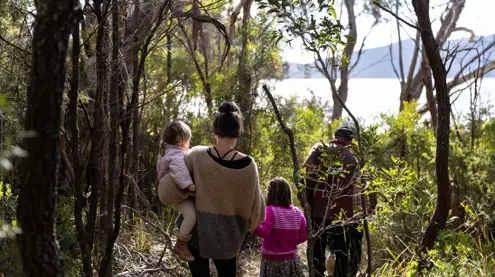 A family walks along a narrow bush track towards the water at Mandala on Bruny Island, with a woman carrying a young child