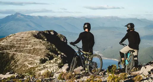 Two mountain bikers on a rocky outcrop overlooking mountains and water at Mount Owen, Tasmania's West Coast