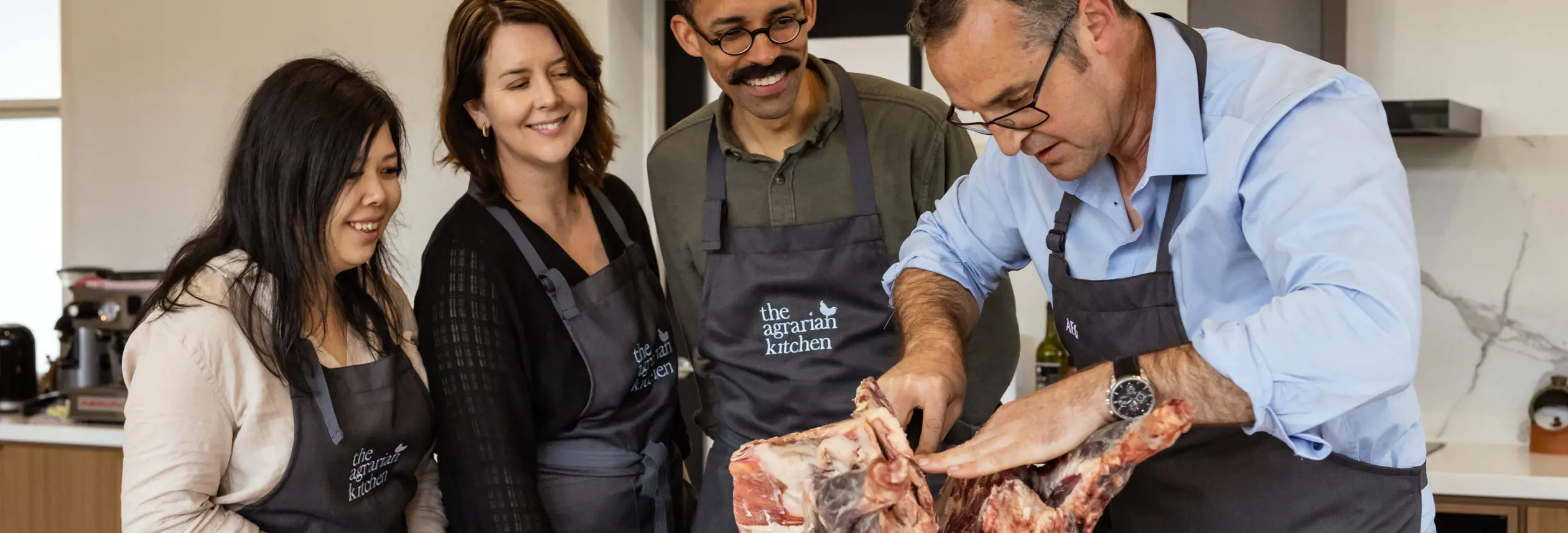 A chef demonstrating butchery to three students in aprons at The Agrarian Kitchen cooking class, New Norfolk