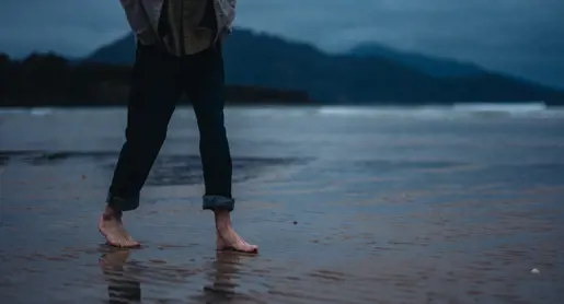A person walks barefoot across wet sand on Sandpiper Beach with mountains and moody blue skies in the background