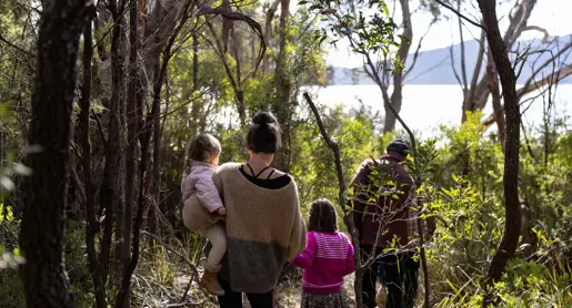 A family walks along a narrow bush track towards the water at Mandala on Bruny Island, with a woman carrying a young child