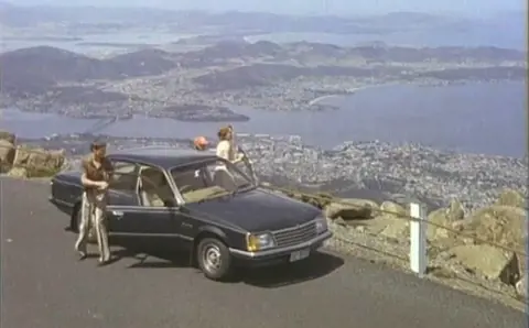 Video screengrab from the 1980 Freewheelin' around Tasmania campaign with travellers and a car overlooking Hobart from kunanyi / Mount Wellington