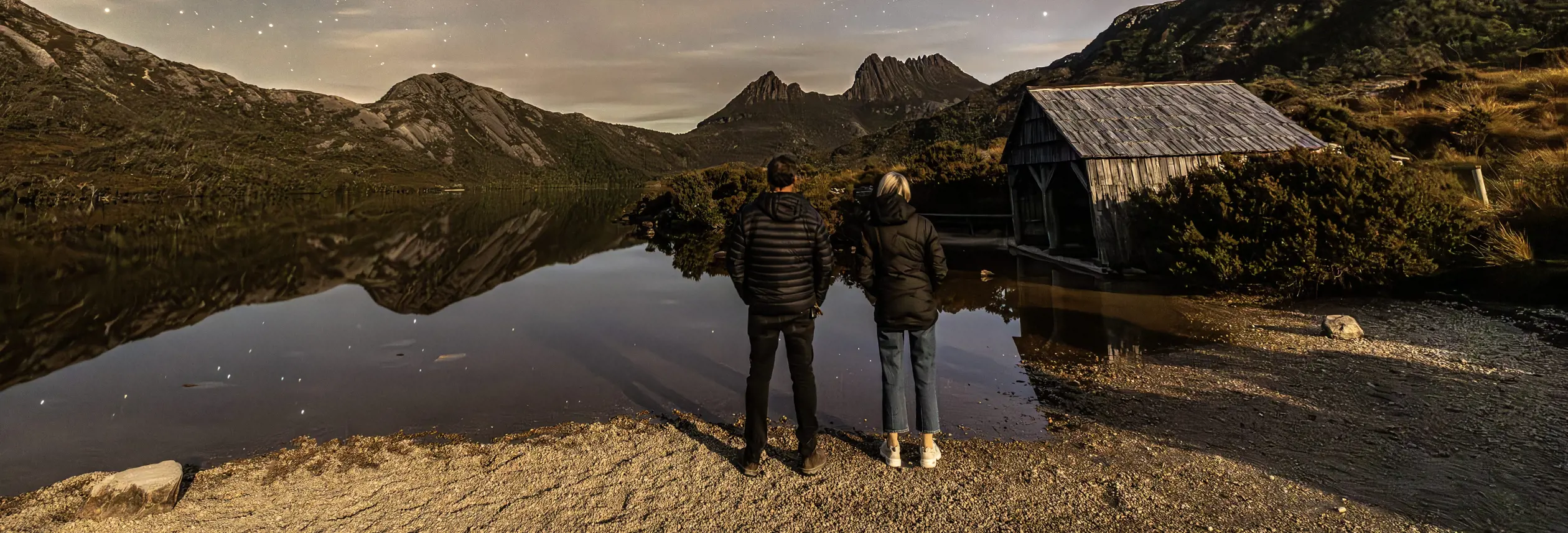 Two people stand at the edge of Dove Lake looking toward Cradle Mountain and a wooden boatshed in warm evening light