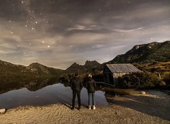Two people stand at the edge of Dove Lake looking toward Cradle Mountain and a wooden boatshed in warm evening light