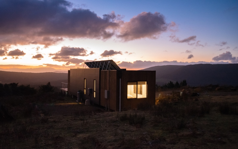 A one-bedroom designer cabin stands on the crest of a hill looking over a valley at dusk.