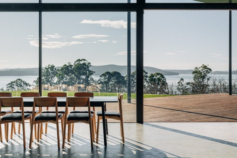 Mid-century style wooden furnture sits on polished concrete floors, looking out over green grass and the D'Entrecasteaux Channel
