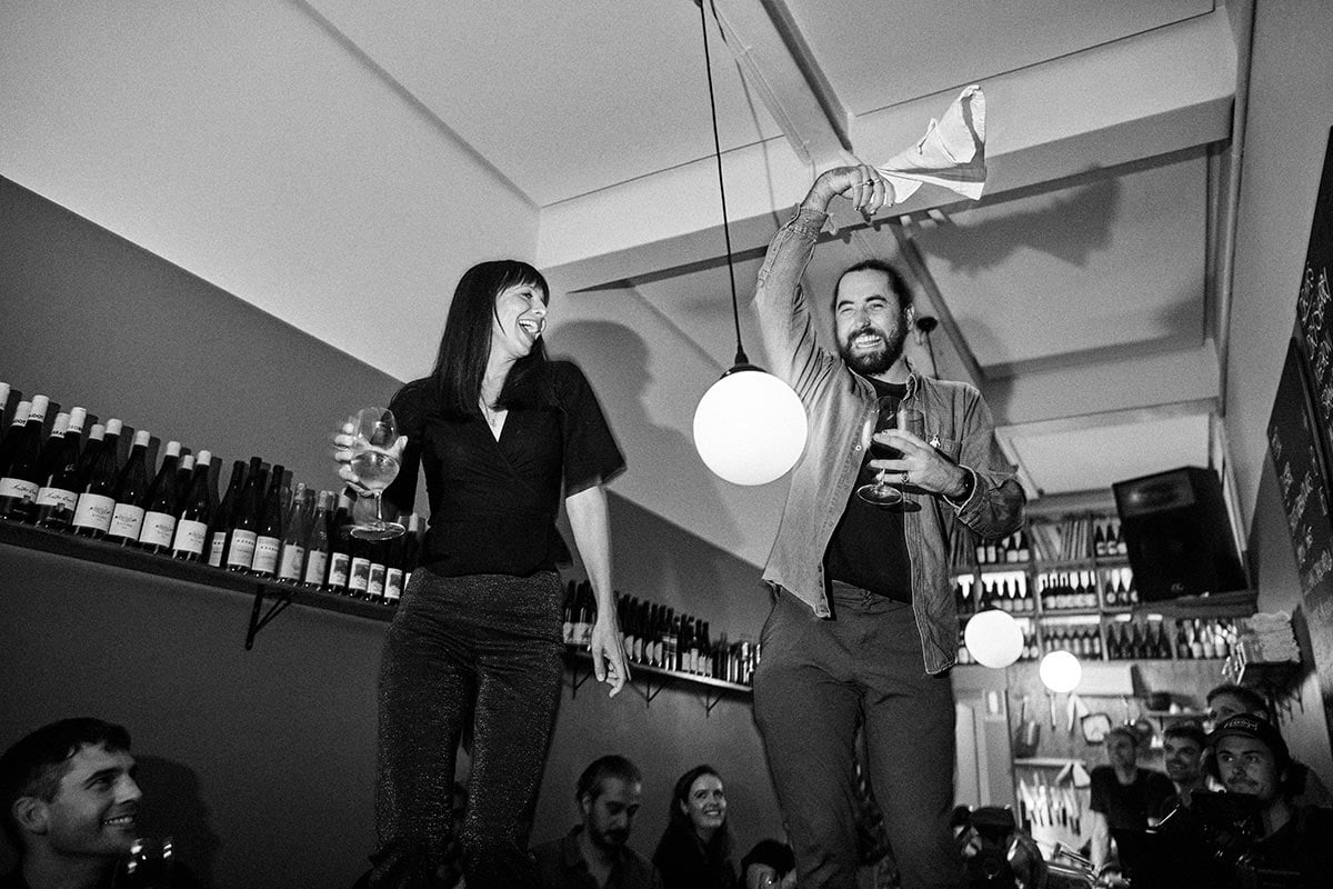 A young woman and young man dance on the top of a bench in a small bar.