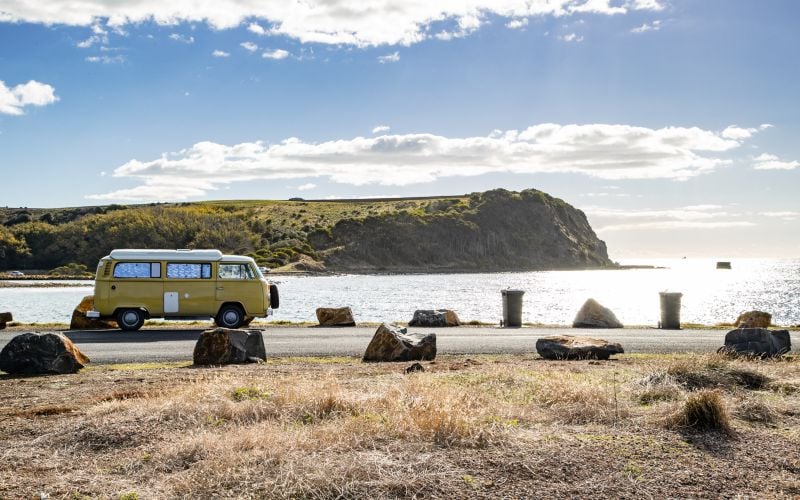 An early model camper van parks at the edge of the road in DEvonport, looking out over the water to a peninsula in the distance