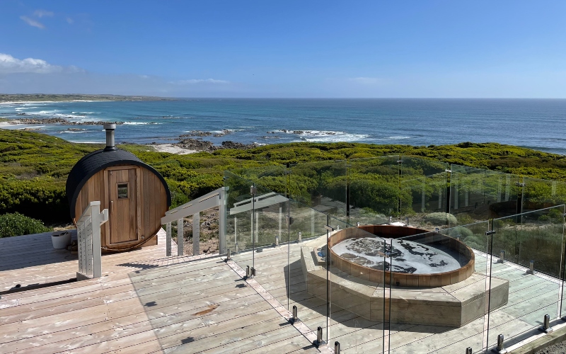 wooden decking with an inset spa bath and glass barriers steps down to a barrel shaped sauna overlooking the beach.